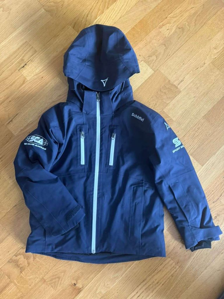 Eine marineblaue Kapuzenjacke mit hellblauen Reißverschlüssen, die flach auf einem Holzboden liegt. Auf den Ärmeln und der Brust sind verschiedene weiße Logos und Texte aufgedruckt, darunter Schlehf und SCA.