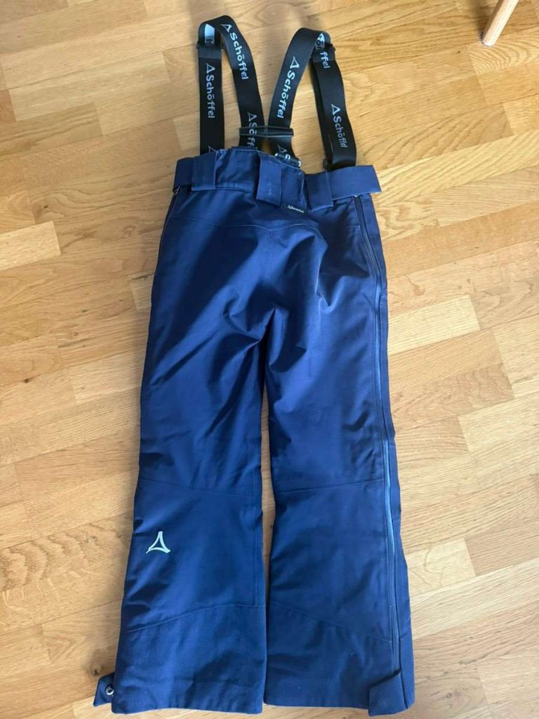 Eine marineblaue Skihose mit schwarzen verstellbaren Hosenträgern, die flach auf einem hellen Holzboden liegt. Die Hose hat einen Gürtel und Reißverschlüsse an den Seiten, mit einem kleinen weißen Logo auf dem linken Bein.